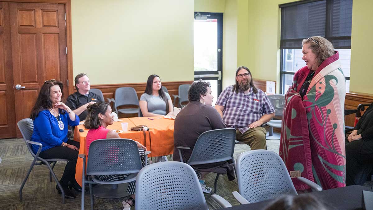 A woman wrapped in a red patterned blanket speaks to a small seated group in a meeting room.