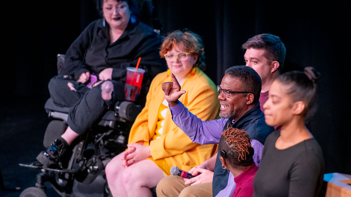 Panelists on stage during a discussion, including a man using sign language and a person in a wheelchair.