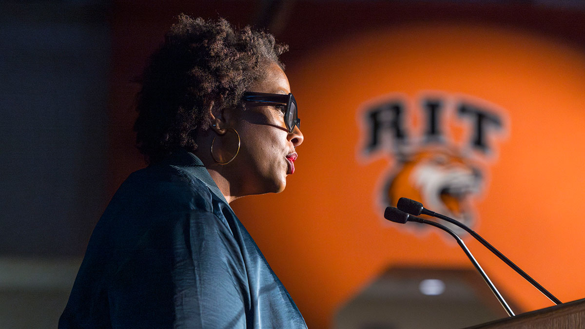 A woman wearing glasses speaks at a podium with an RIT Tigers logo in the background.