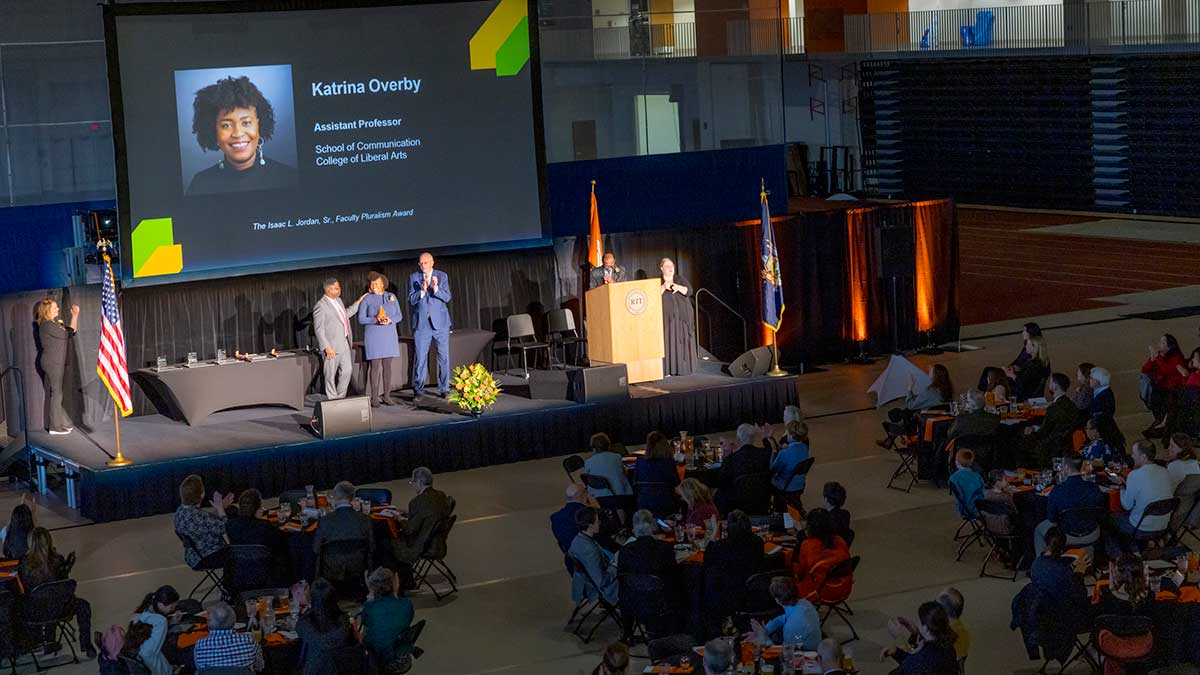 A stage presentation honors Katrina Overby with an award as attendees watch from banquet tables.
