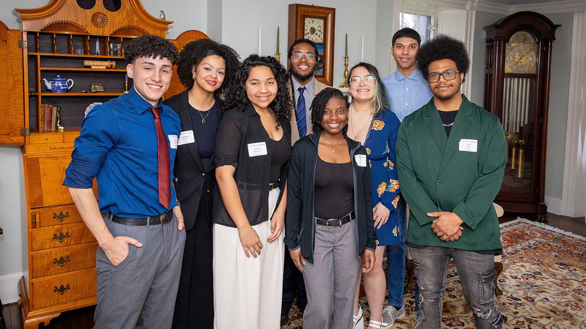 Eight students pose together indoors, smiling in front of antique wooden furniture.