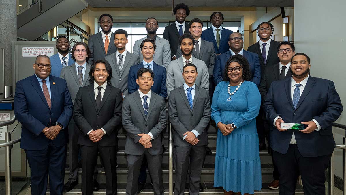 18 men in suits and a woman in a blue dress pose together on a staircase inside a modern building.