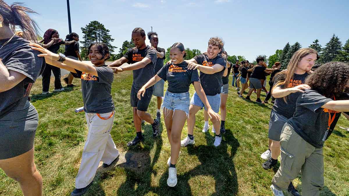Students participate in an outdoor conga line during a summer program activity on a sunny day.