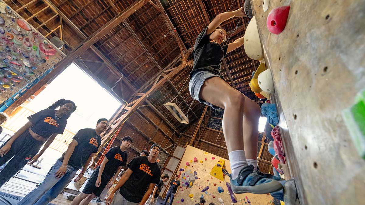 A young woman climbs an indoor rock wall while other students watch during a summer program.
