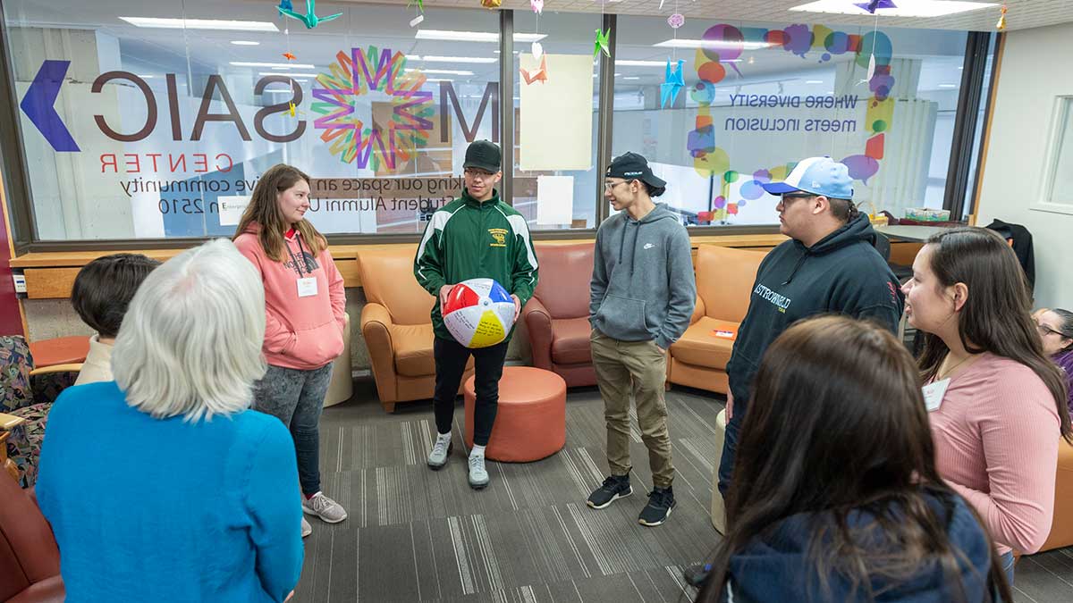 students standing in a circle in a room, with one student holding a beach ball with writing on it.