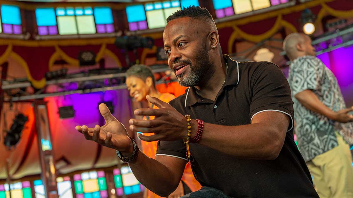A man gestures expressively while speaking on stage with colorful stained glass panels in the background.