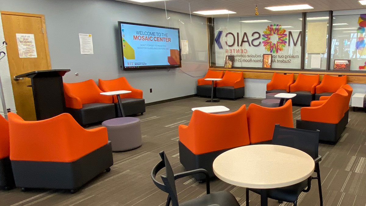A lounge with orange and black chairs and a screen welcomes visitors to the RIT Mosaic Center.