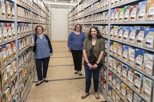 Three women standing in archiche shelving rows