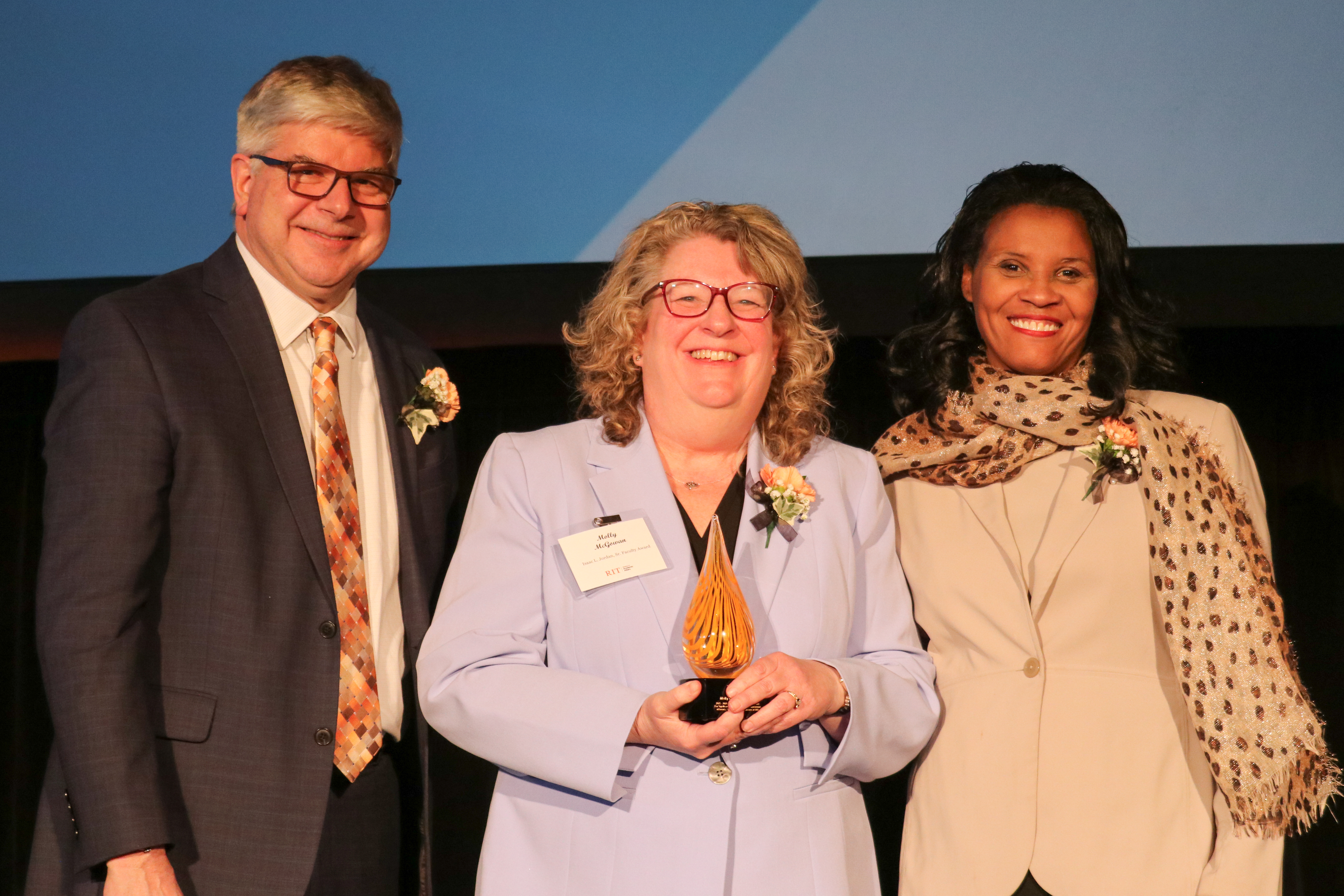 Three people posing on stage. Middle woman is holding an award