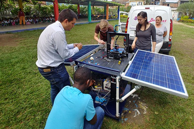 RIT students and Professor Rojas are outside setting up their solar powered 3D printer. In the distance, a large group of school children look at their progress.