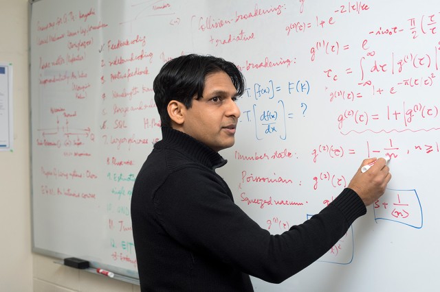 Mishkat Bhattacharya standing in front of white board and writing out equations.