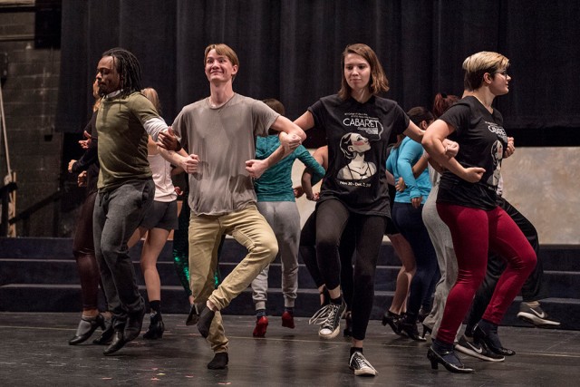 Members of the cast practice a group dance, their arms linked together as they move around in a circle.
