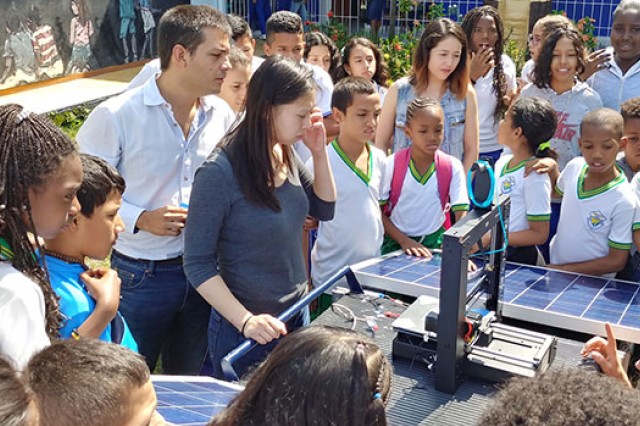 Team members demonstrate their new 3D printer and young students crowd around them to look and see the machine function.