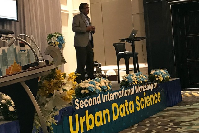 A man stands on stage and talks to a group of people. Attached to the edge of the stage is a banner that reads "Second International Workshop on Urban Data Science." There are also several flower arrangements scattered across the stage.