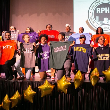 A group of graduating students from Rochester Prep High School stand on a stage and hold up t-shirts from the colleges they have been accepted into for the fall.