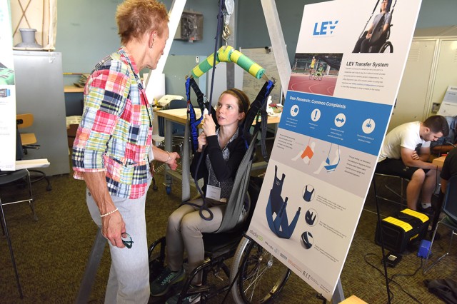 Ana Sorensen sits in a wheelchair and demonstrates how the LEV lift works for individuals to lift themselves up. She shows this to another woman who is engaged in the demonstration and reaches out to touch the assistive device.