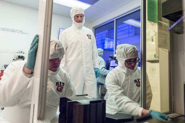 Three researchers dressed in white, sterile jumpsuits work together in a lab.
