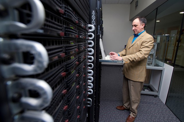 Man in tan suit stands and types on keyboard of supercomputer