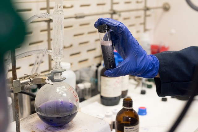 Gloved hand hold testtube of black liquid; container with purple liquid in background