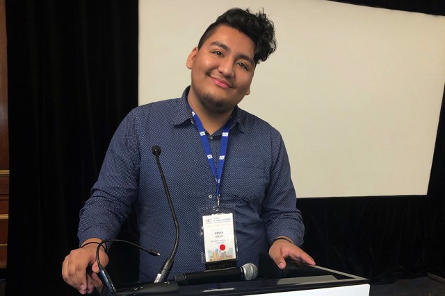 Student wearing blue button-up shirt and name tag stands at podium.