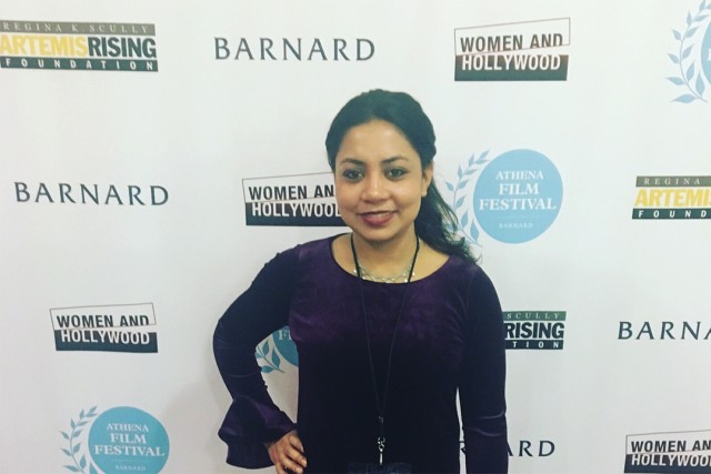 Woman in purple shirt standing in front of white background with logos.