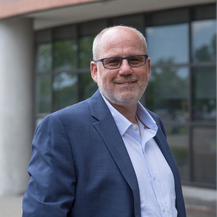 Man wearing glasses and blue suit stands outside of building.