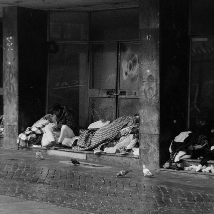 Homeless people with blankets near doors of building.