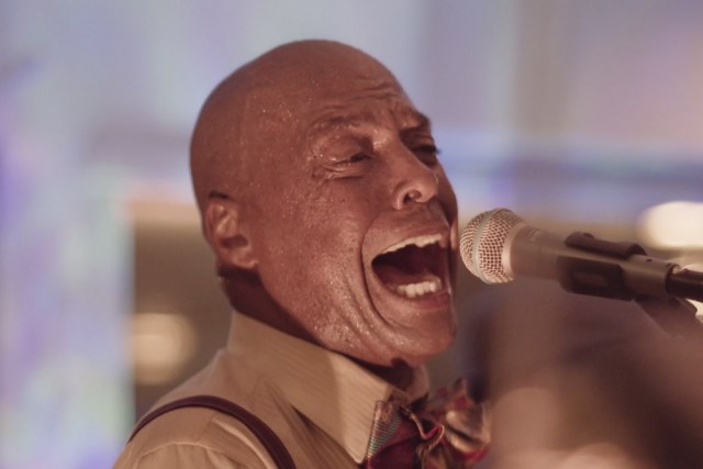 screengrab from a film in which a man is singing into a microphone.