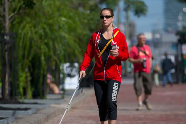 person walking wearing sunglasses, holding a smartphone, and using a guide cane.