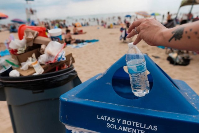 Hand puts plastic water bottle in a blue recycling in on a busy beach.