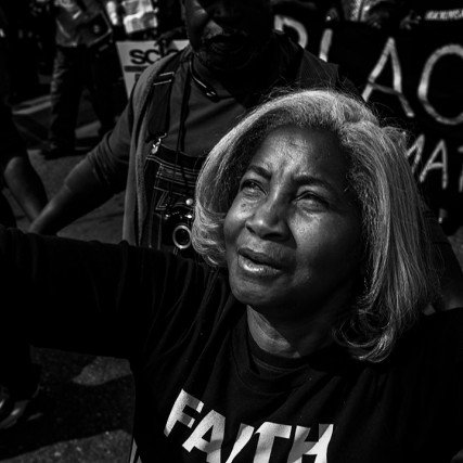 black and white photo of a woman with her arms outstretched and a Black Lives Matter sign in the background.