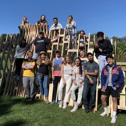A group of RIT designers and architects post for a photo on the temporary wood installation they created on the grounds of the 1969 Woodstock festival.