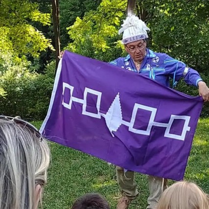 person dress in traditional Native American clothing holding a purple flag.