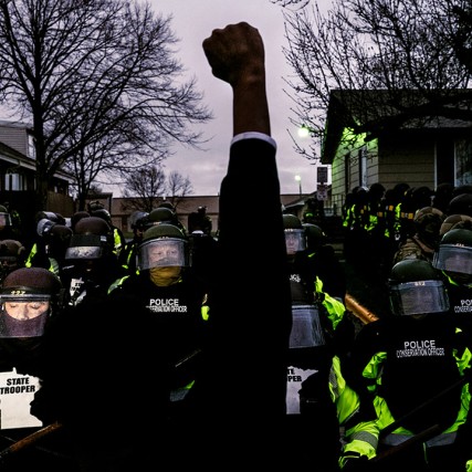 a person raising a fist in the air in front of riot control police officers.