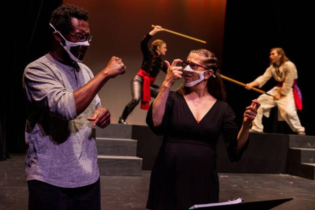 two people using sign language in the foreground and actors in a play on a stage in the background.