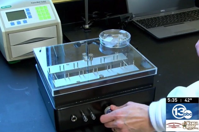 screenshot from a news report featuring a hand turning a knob on a rectangular device with a clear top.