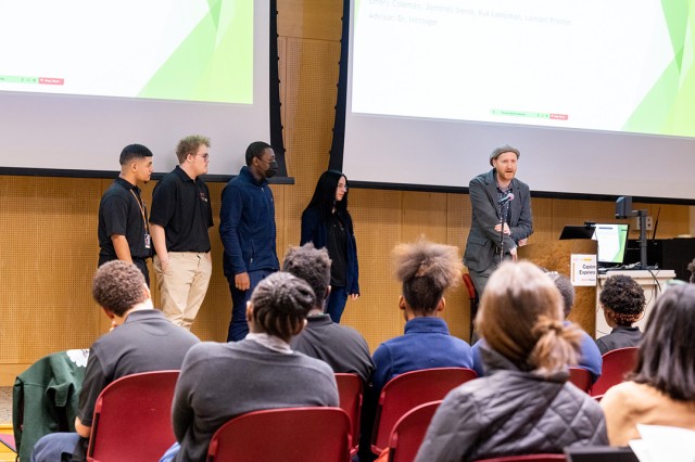 four students on a stage and a professor talking into a microphone.
