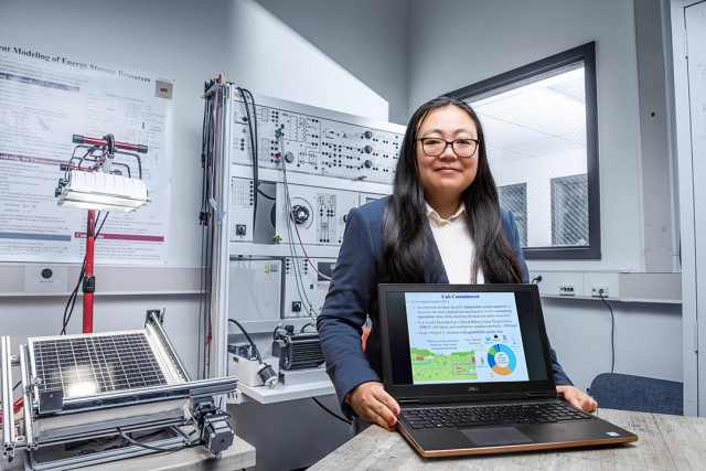 Bing Yan is shown working in a lab holding a laptop that is facing the camera showing a slide from a presentation deck