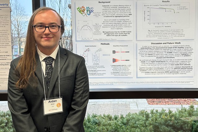 Aiden Miller is shown posing in front of their research which is hanging on a building window.