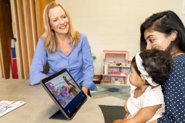 Rain Bosworth smiling and looking at a parent-child pair to her left. She has blonde hair and blue eyes and wearing blue button-up shirt. The parent is looking at an iPad, sitting in front of them on a round table. The iPad is displaying what appears to be a video with a person signing. The parent has black hair and wearing a navy polka dot shirt. The child is sitting on the parent's lap and staring at Bosworth.