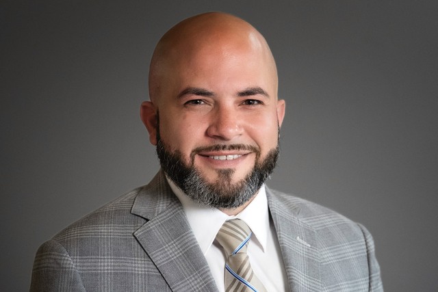 Orlando Ortiz sits for a headshot against a gray background wearing a gray plaid suit.
