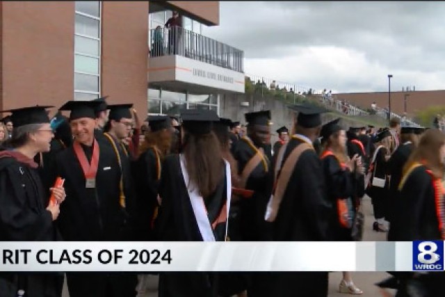 a crowd of graduates in regalia are shown standing in front of the Gordon Field House.