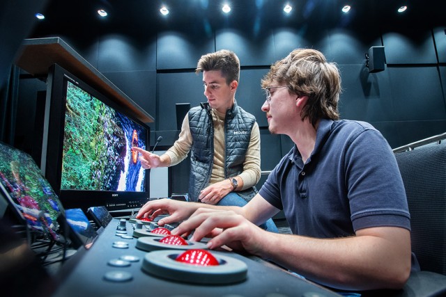 two students stand in front of a small screen reviewing footage in the MAGIC Center Color Correction Suite on campus.