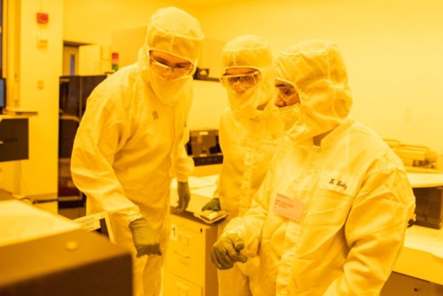 Three people in a yellow-tinted clean room wearing white clean suits, gloves, and safety glasses.