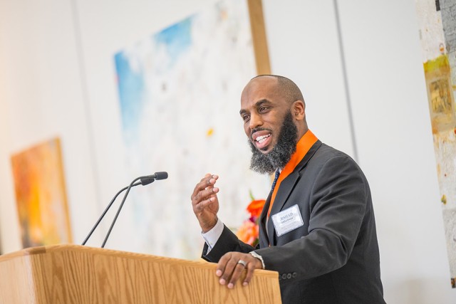a black man with a beard stands at a podium wearing a black suit jacket with an orange dress shirt, addressing an audience.