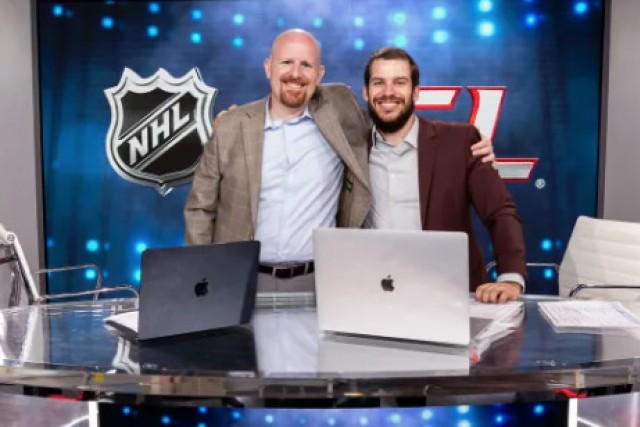 Broadcasters Noah Blankenship and Jason Altmann stand in front of an NHL sign with a small table in front of them.