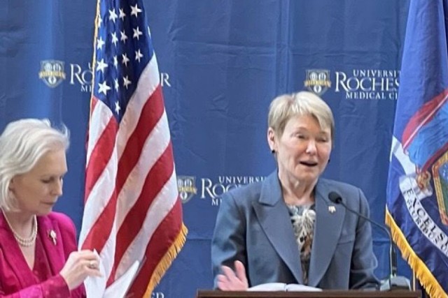 Two women stand at a podium during an event at the University of Rochester Medical Center. One woman speaks into a microphone while gesturing, and the other adjusts an American flag. A backdrop displays the university's logo.