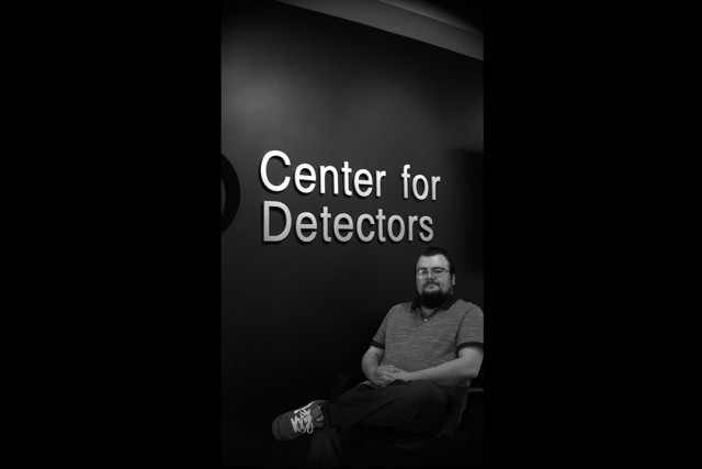 A bearded man wearing glasses sits in a chair in front of a black wall with 'Center for Detectors' in large, bold lettering.