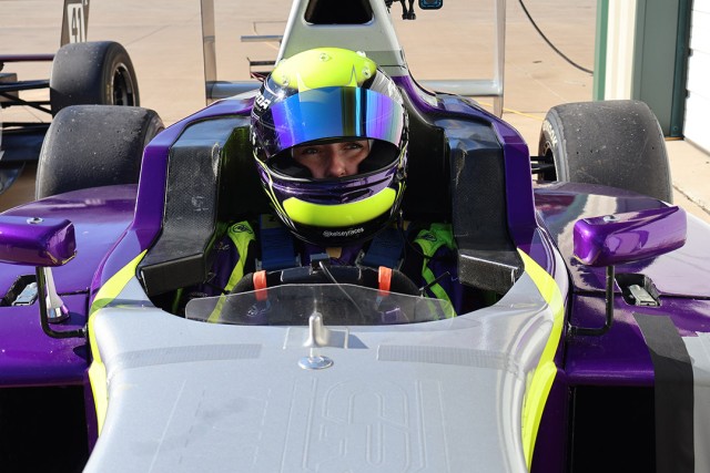 Race car driver wearing a vibrant yellow and purple helmet sits inside a purple open-wheel race car, gripping the wheel with a focused expression.