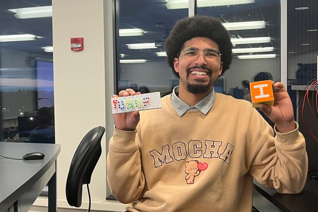 Smiling man with glasses and an afro hairstyle holds up a breadboard with electronic components in one hand and a small orange and white 3D-printed block with the letter 'I' in the other. He wears a beige sweater with 'MOCHA' and a bear design, sitting in a modern classroom or lab setting with computers and wiring in the background.